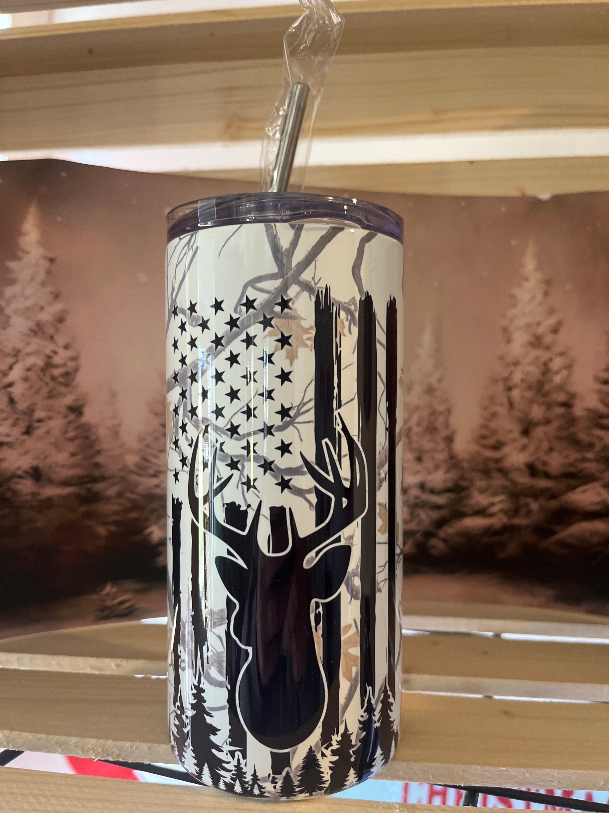A 16 oz tumbler with a camouflage pattern and an illustration of a deer's head and antlers, alongside an American flag, set against a backdrop of a forest.