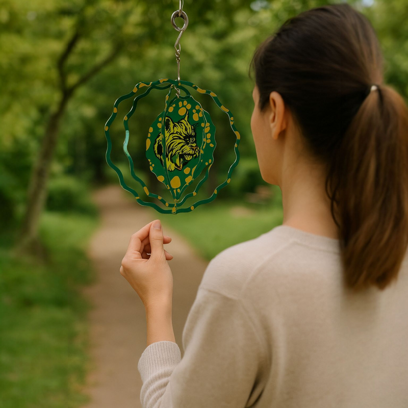 A person standing in an outdoor setting holding a green metal wind spinner with Florence Bobcat logo pattern.